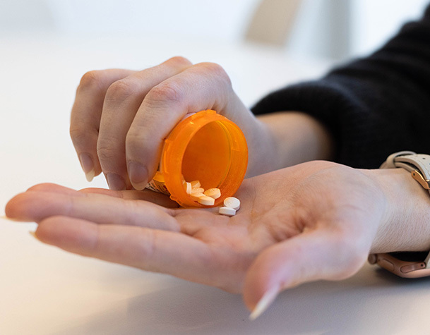 The image shows a person holding an orange prescription pill bottle, pouring white pills into their open hand. 