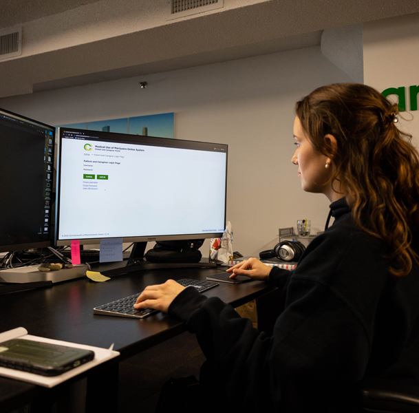 The image shows a jd esoftware employee sitting at a desk, working on a computer with a large monitor displaying the Medical Use of Marijuana Online System.